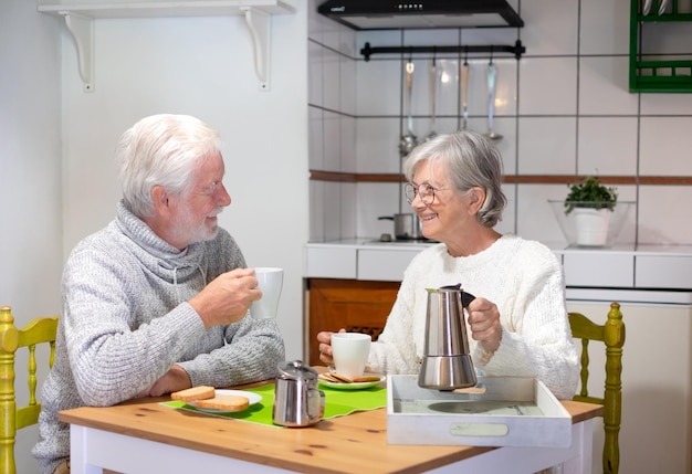 Glimlachend senior paar samen ontbijten aan huistafel, bejaarde vrouw met koffiezetapparaat