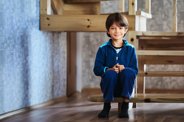 Glimlachend slank Europees mannelijk kind gekleed in sportuniform zit op de trap van een houten trap die leidt