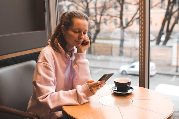 Glimlachend stijlvol jong meisje 22-24 jaar oud bericht aan het typen op telefoon koffie drinken in café close-up.