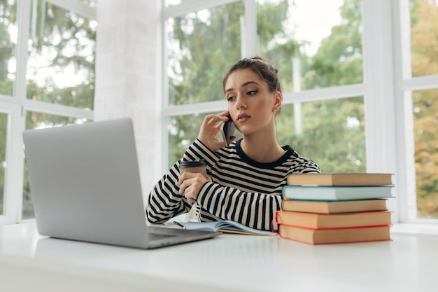 Glimlachend student meisje met laptop praten op mobiele telefoon zittend aan de balie thuis