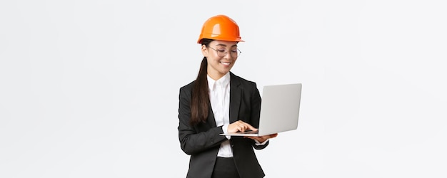 Glimlachend succesvolle vrouwelijke Aziatische industrieel ingenieur fabrieksmanager in veiligheidshelm en pak met behulp van laptopcomputer controleren met project of blauwdrukken op het scherm