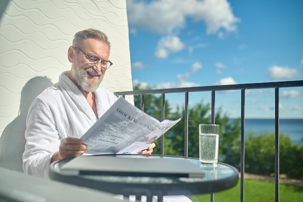 Glimlachend tevreden man in bril en de witte badjas zittend aan de glazen salontafel op het balkon