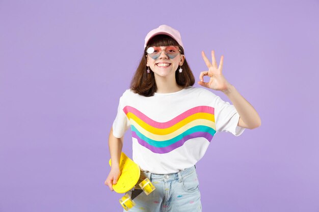 Glimlachend tienermeisje in levendige kleding, hartbrillen houden geel skateboard, met OK gebaar geïsoleerd op violet pastel muur achtergrond. Mensen oprechte emoties, lifestyle concept. Bespotten kopie ruimte