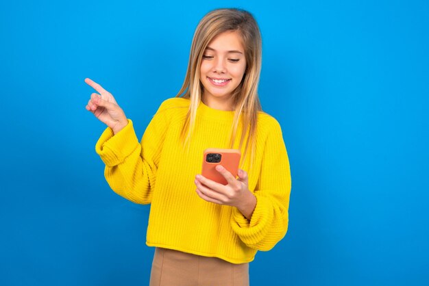 Glimlachend tienermeisje met gele trui wijzende vinger naar lege ruimte met telefoon in één hand