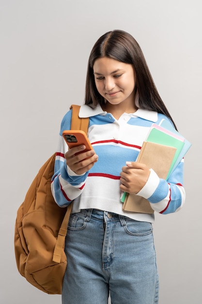 Glimlachend tienermeisje met rugzak kijkt naar het scherm van zijn smartphone op een grijze achtergrond