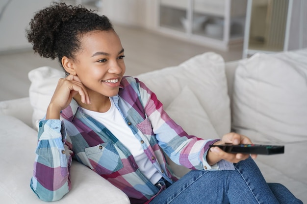 Glimlachend tienermeisje van gemengd ras dat naar televisieseries kijkt, verandert van tv-kanaal terwijl ze thuis op de bank zit