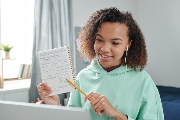 Glimlachend tienermeisje van gemengd ras met oortelefoon wijzend op notities op pagina van beurt terwijl ze ze uitlegt aan haar vriend op laptopscherm