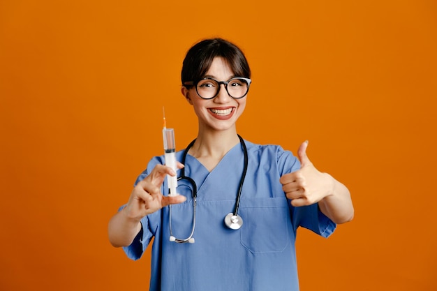 Glimlachend tonen duimen omhoog houden spuit jonge vrouwelijke arts dragen uniform fith stethoscoop geïsoleerd op oranje achtergrond