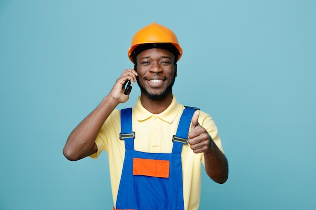Glimlachend tonen duimen omhoog jonge Afro-Amerikaanse bouwer in uniform spreekt aan de telefoon geïsoleerd op blauwe achtergrond