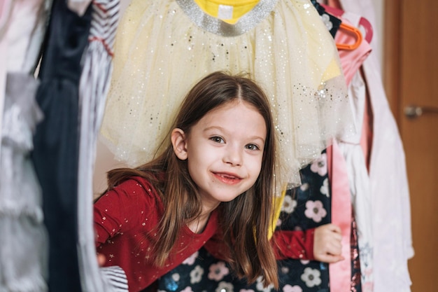 Glimlachend tween meisje met lang donker haar in rode jurk tussen haar prachtige jurken in kledingkast in de kinderkamer thuis