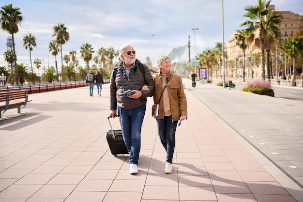 Glimlachend volwassen blank toeristisch echtpaar loopt omhelsd zorgeloos met bagage op een zonnige dag buiten