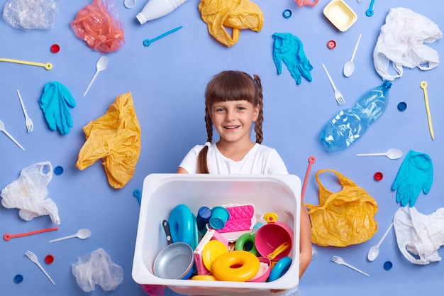 Glimlachend vriendelijk meisje met een wit t-shirt poserend tegen een blauwe muur en veel plastic afval in de buurt weigert van al haar plastic speelgoed voor de bescherming van de aarde