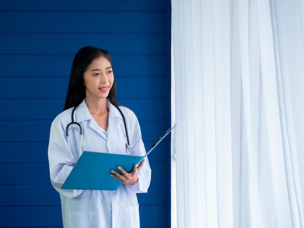 Glimlachend vrij Aziatische vrouw arts portret staande op blauwe houten en witte gordijn achtergrond in ziekenhuis kamer Zelfverzekerde Aziatische jonge vrouwelijke beoefenaar met stethoscoop document map te houden