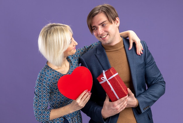 Glimlachend vrij jong stel dat naar elkaar kijkt met een geschenkdoos en een rode hartvorm op valentijnsdag