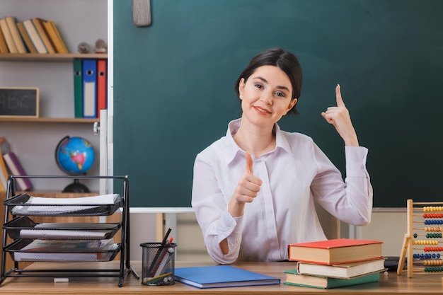 glimlachend wijst omhoog met duimen omhoog jonge vrouwelijke leraar zit aan bureau met schoolhulpmiddelen in de klas