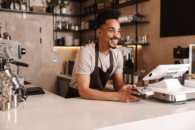 Glimlachende aantrekkelijke Afrikaanse man barista staande achter de toonbank in de coffeeshop