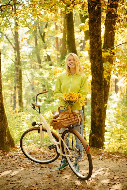 Glimlachende aantrekkelijke jonge vrouw in herfstpark met bloemen in de buurt van haar retro fiets Blonde en