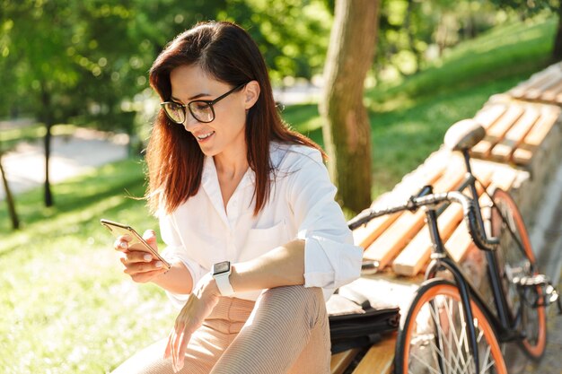 Glimlachende aantrekkelijke jonge vrouw ontspannen op de bank in het stadspark, met behulp van mobiele telefoon