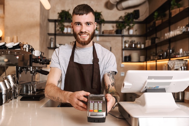 Glimlachende aantrekkelijke man-barista die achter de toonbank in de coffeeshop staat en kaartlezerapparaat geeft