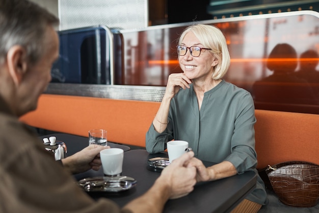 Glimlachende aantrekkelijke senior blanke vrouw in bril zittend aan tafel in bistro café en hand van vriendje vasthoudend terwijl hij naar hem luistert