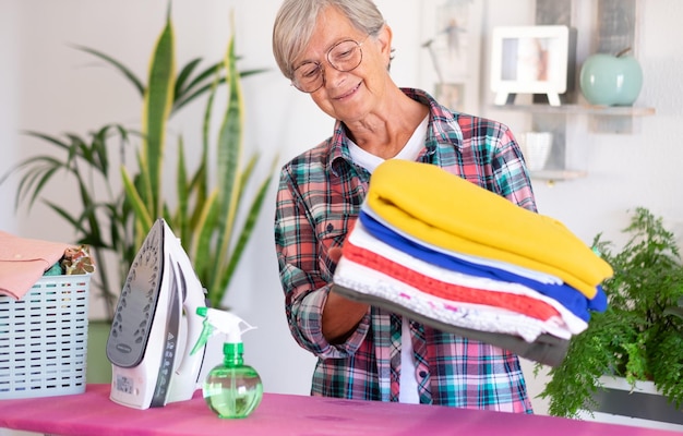 Glimlachende aantrekkelijke senior vrouw in geruit hemd en bril strijken kleren thuis op strijkplank