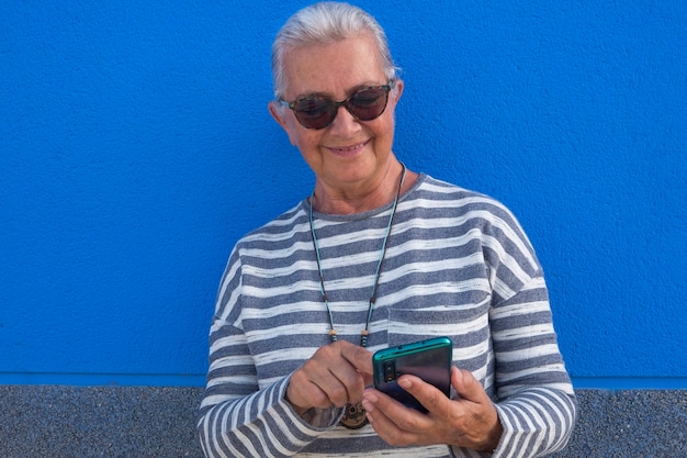 Glimlachende aantrekkelijke senior vrouw met wit haar met behulp van slimme telefoon app een bericht sturen naar vrienden - blauwe achtergrond - tech en sociale ouderen