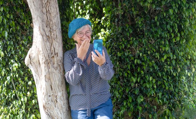 Glimlachende aantrekkelijke senior vrouw zittend in de tuin onder een boom met behulp van mobiele telefoon in video-oproep Groene achtergrond