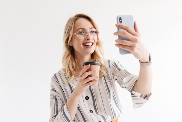 Glimlachende aantrekkelijke vrouw met een bril die een selfie-foto neemt en een papieren beker vasthoudt die over een witte muur wordt geïsoleerd
