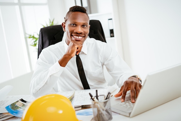 Glimlachende Afrikaanse bouwingenieur zit aan de balie op kantoor en kijkt naar de camera.