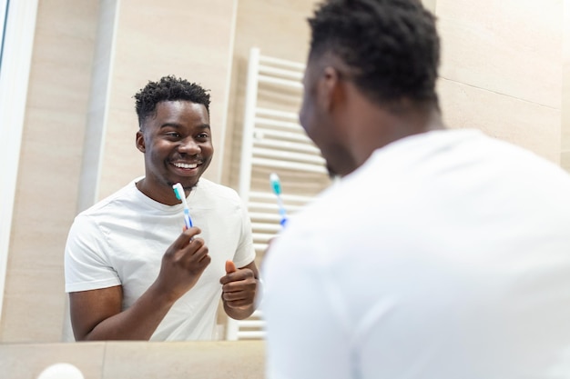 Glimlachende Afrikaanse man met tandenborstel die tanden schoonmaakt en spiegel in de badkamer kijkt