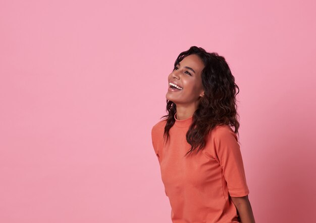 Glimlachende Afrikaanse vrouw die exemplaarruimte bekijkt die toevallige oranje die t-shirt draagt op roze wordt geïsoleerd.