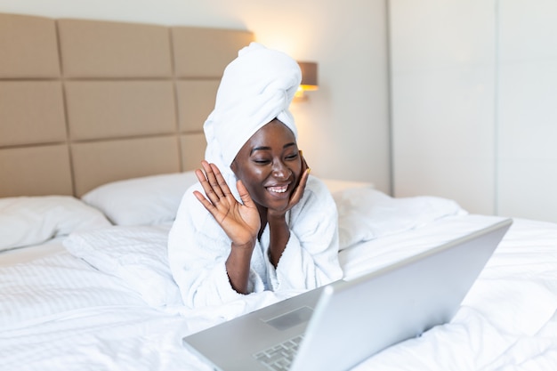Glimlachende Afrikaanse vrouw die op bed in badjas met laptop ligt die met haar vrienden via videogesprek spreekt.