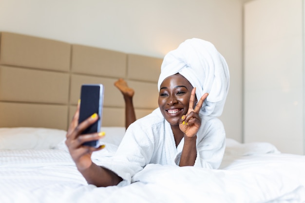 Glimlachende Afrikaanse vrouw die op bed in badjas met mobiele telefoon ligt die een selfie neemt. vredesteken tonen