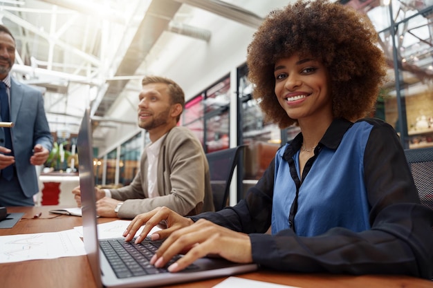 Glimlachende Afrikaanse zakenvrouw zittend op zijn werkplek in modern kantoor op collega's achtergrond