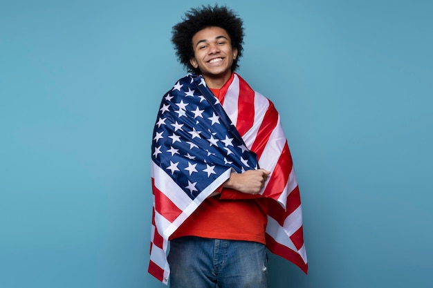 Glimlachende Afro-Amerikaanse man die bedekt is met Amerikaanse vlag geïsoleerd op blauwe achtergrond. 4 juli