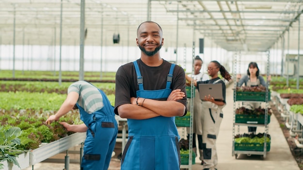 Glimlachende Afro-Amerikaanse man poseren met gekruiste armen terwijl landarbeiders met behulp van laptop leveringen aan lokale winkel beheren. Portret van een boer die in een moderne kas staat terwijl plukkers kratten vullen.