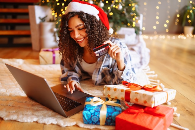 Glimlachende Afro-Amerikaanse vrouw die een laptop gebruikt die in de buurt van de kerstboom zit met een kaart die online winkelt