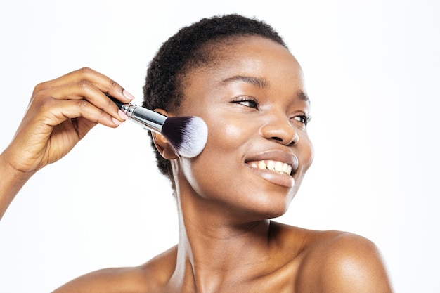 Glimlachende Afro-Amerikaanse vrouw die make-up aanbrengt met borstel geïsoleerd op een witte achtergrond white