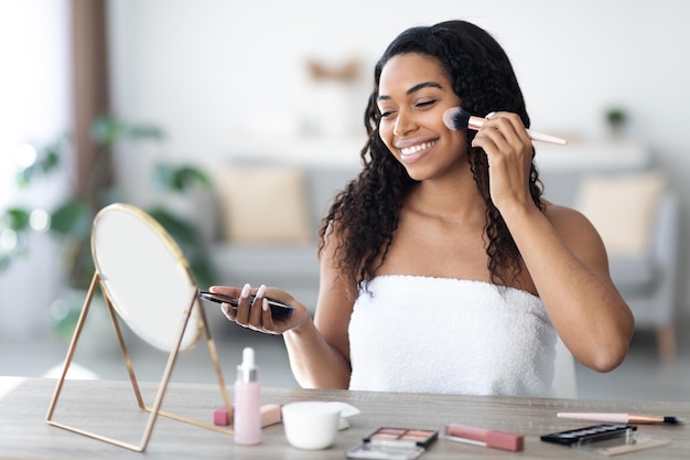 Glimlachende Afro-Amerikaanse vrouw gewikkeld in een handdoek die make-up opdoet