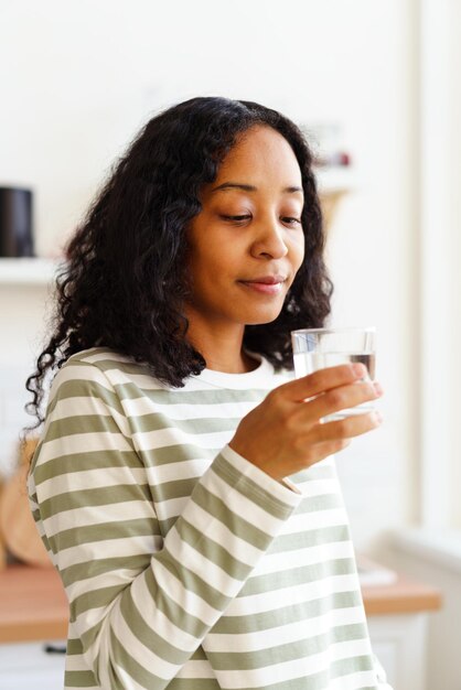 Foto glimlachende afro-amerikaanse vrouw kijkt naar een glas helder water op een zachte, gefocuste achtergrond.