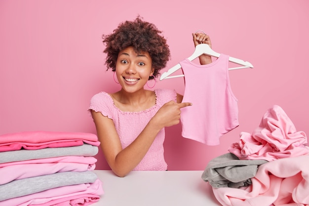 Glimlachende Afro-Amerikaanse vrouw wijst naar haar t-shirt op hanger sorteert kleding voor donatie omringd door stapel ongevouwen wasgoed stapel netjes opgevouwen kleding poses binnen tegen roze muur