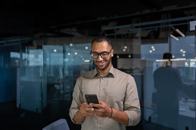 Glimlachende Afro-Amerikaanse zakenman in kantoor in shirt met behulp van telefoonman die bericht typt en