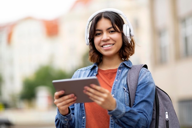 Glimlachende Arabische vrouwelijke student in draadloze koptelefoon met behulp van digitale tablet buitenshuis