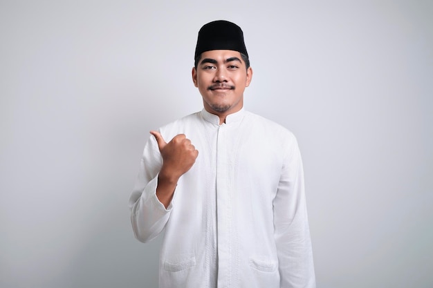 Glimlachende aziatische jonge moslimman in witte kleding en schedelkap wijzend naar linksboven om ruimte te kopiëren met zijn handvinger