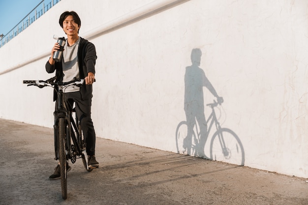 Glimlachende aziatische man die buiten fietst, drinkt water uit een fles