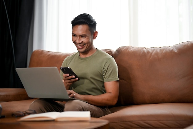 Glimlachende Aziatische man die zijn smartphone en laptop gebruikt terwijl hij op een bank in zijn woonkamer zit