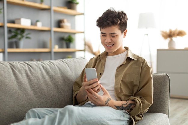 Glimlachende Aziatische man met behulp van telefoon thuis zittend op de bank