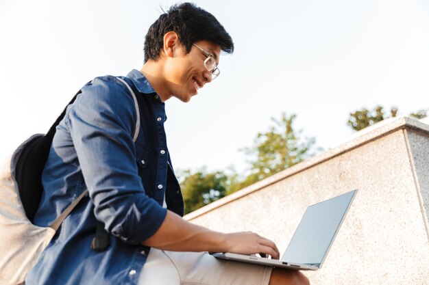 Glimlachende Aziatische man student met rugzak met laptop zittend buiten