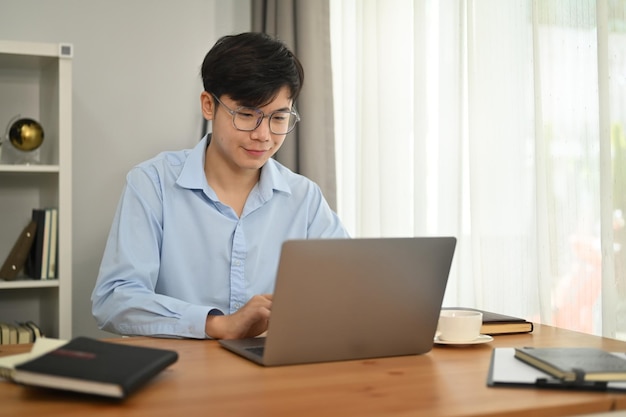 Glimlachende Aziatische mannelijke freelancer in brillen die op afstand werken aan online project op laptop in helder kantoor aan huis