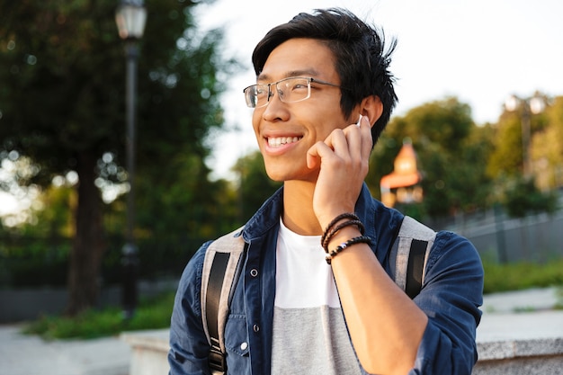 Glimlachende Aziatische mannelijke student in oogglazen die door smartphone spreken en weg buiten kijken